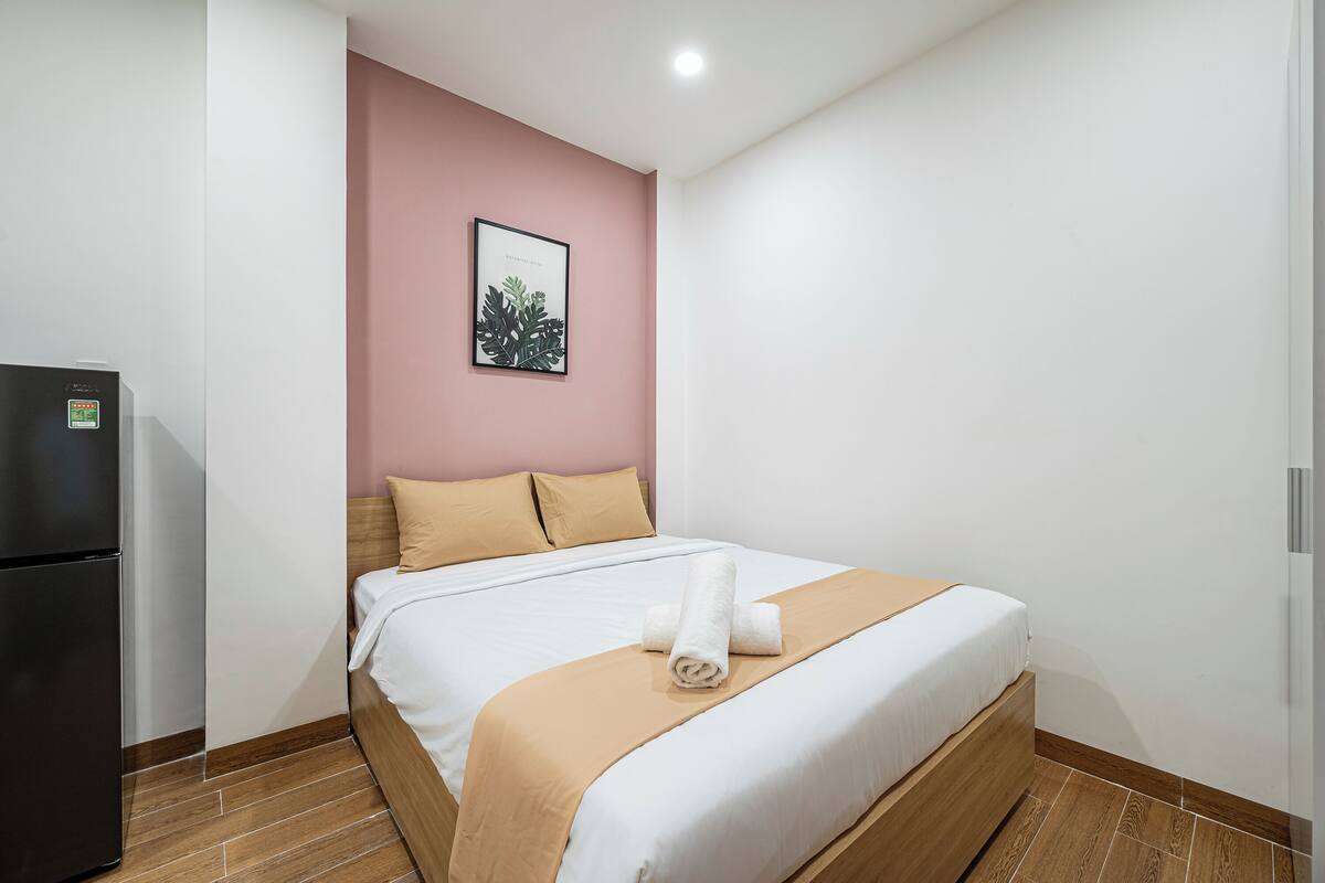 Una cama bien tendida con una colcha blanca y beige, un cabecero marrón y una toalla enrollada sobre la cama.