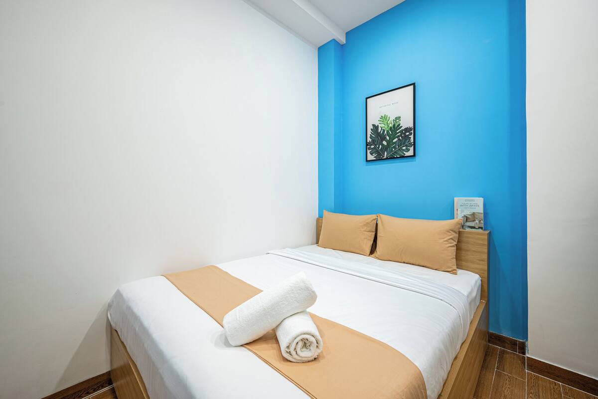 Una cama bien tendida con una colcha blanca y beige, una toalla blanca enrollada y un cojín color arena.