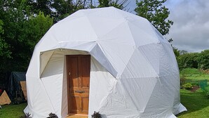 Uma tenda em forma de abóbada com porta de madeira, cercada por uma área gramada e flores coloridas.
