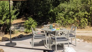 An outdoor patio with a table and chairs, a table umbrella, and a view of a forested area. - Clairette- Charming gîte, 2 bedrooms (Gonfaron)