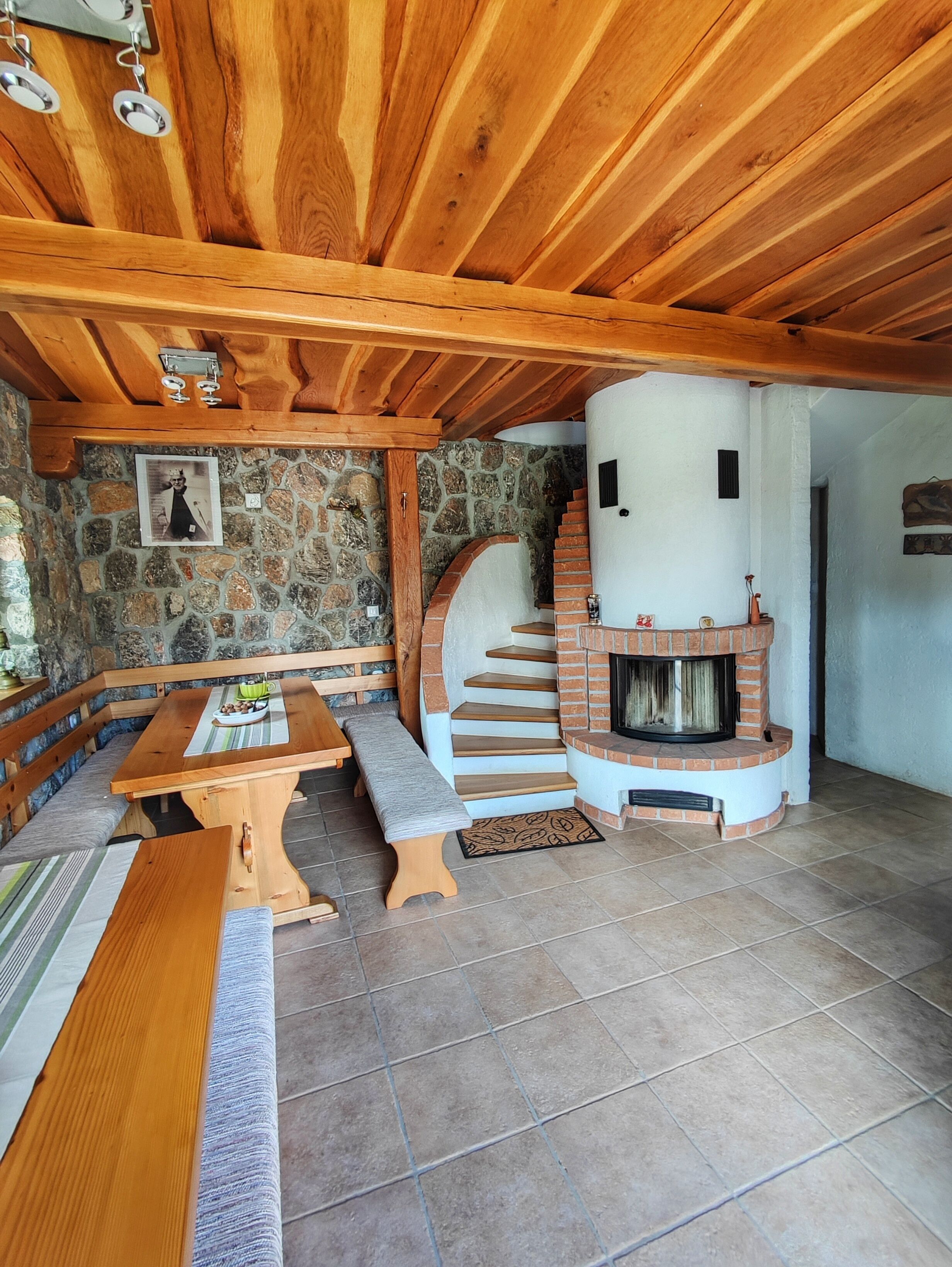 Ein rustikaler Innenraum mit einer Steinwand, einer hölzernen Bank, einem Kamin und einer Treppe, die zu einer oberen Ebene führt.