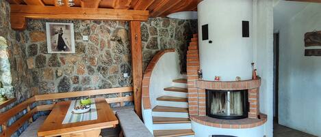 Ein rustikaler Innenraum mit einer Steinwand, einer hölzernen Bank, einem Kamin und einer Treppe, die zu einer oberen Ebene führt.
