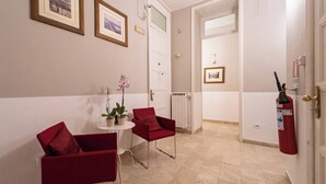 Een hotelkamer met twee rode stoelen, een kleine tafel met een potplant en een brandblusser aan de muur.
