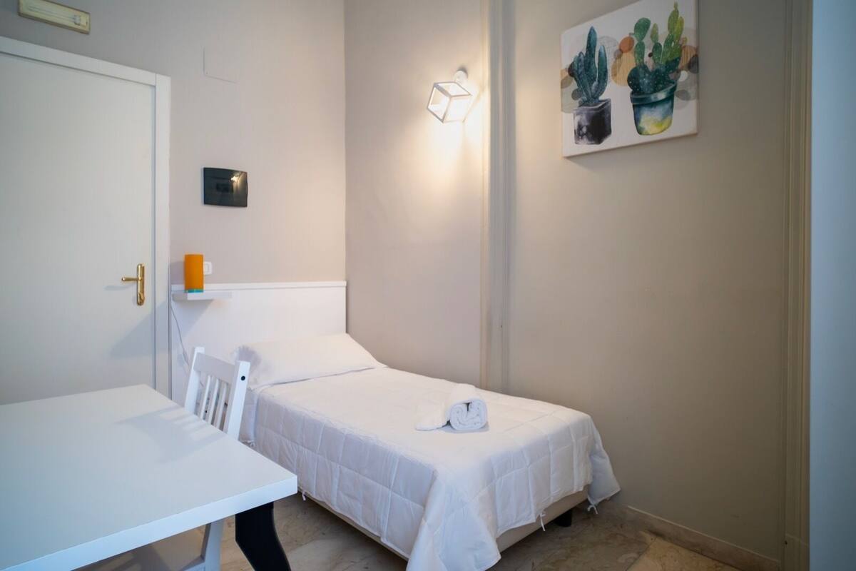 A small, neatly arranged room with a single bed, a white table, and a wall-mounted light fixture.