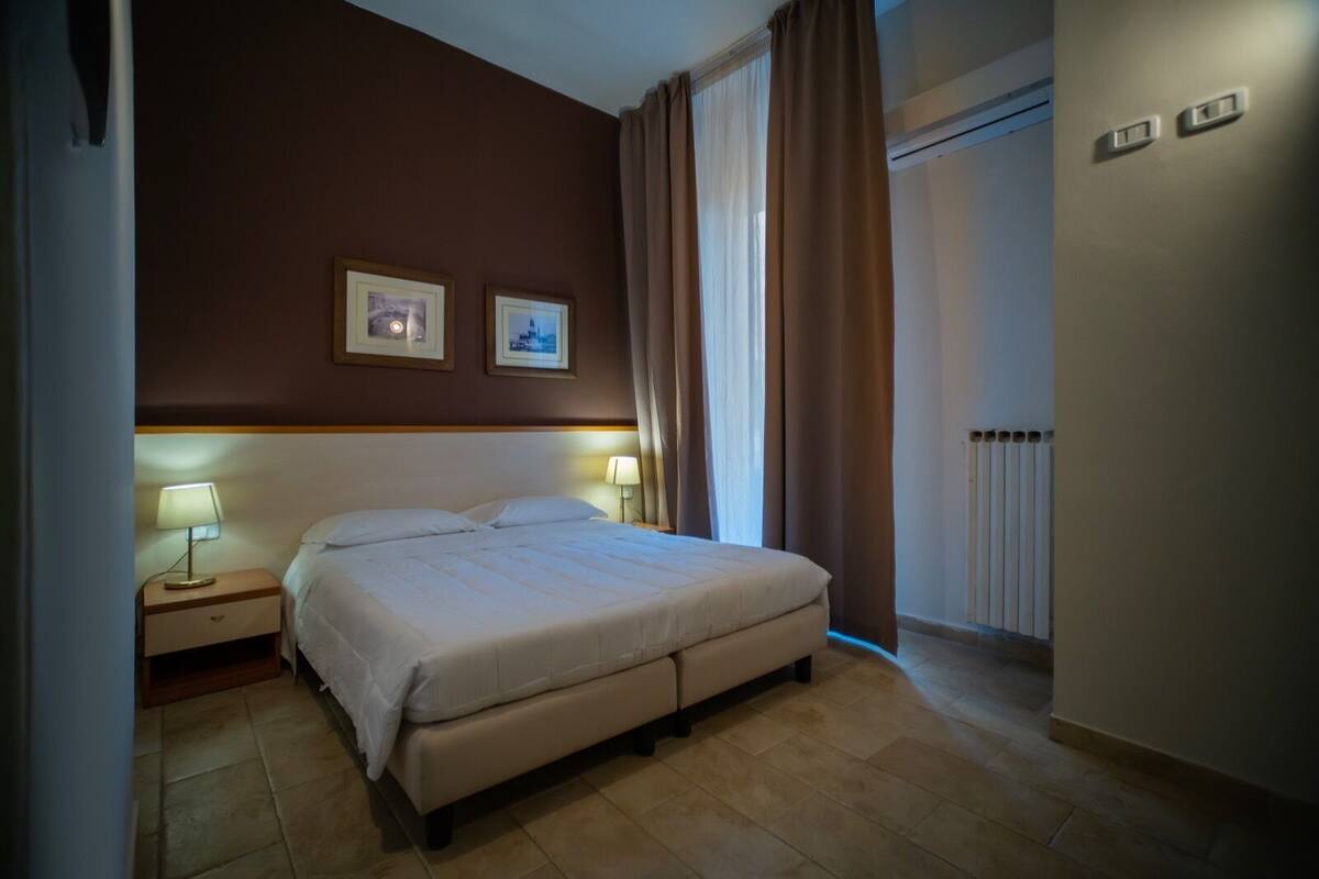 A hotel room with a bed, two bedside lamps, a radiator, and two framed pictures on the wall.