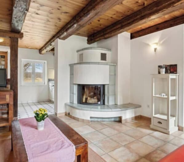 Una habitación con chimenea, techo de madera, suelo de baldosas y una estantería blanca.