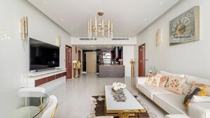 Ein geräumiges Wohnzimmer mit einem großen Flachbildfernseher, einer Kristallleuchter, einem Sofa mit floralen Kissen und einem Essbereich mit einem Glastisch.