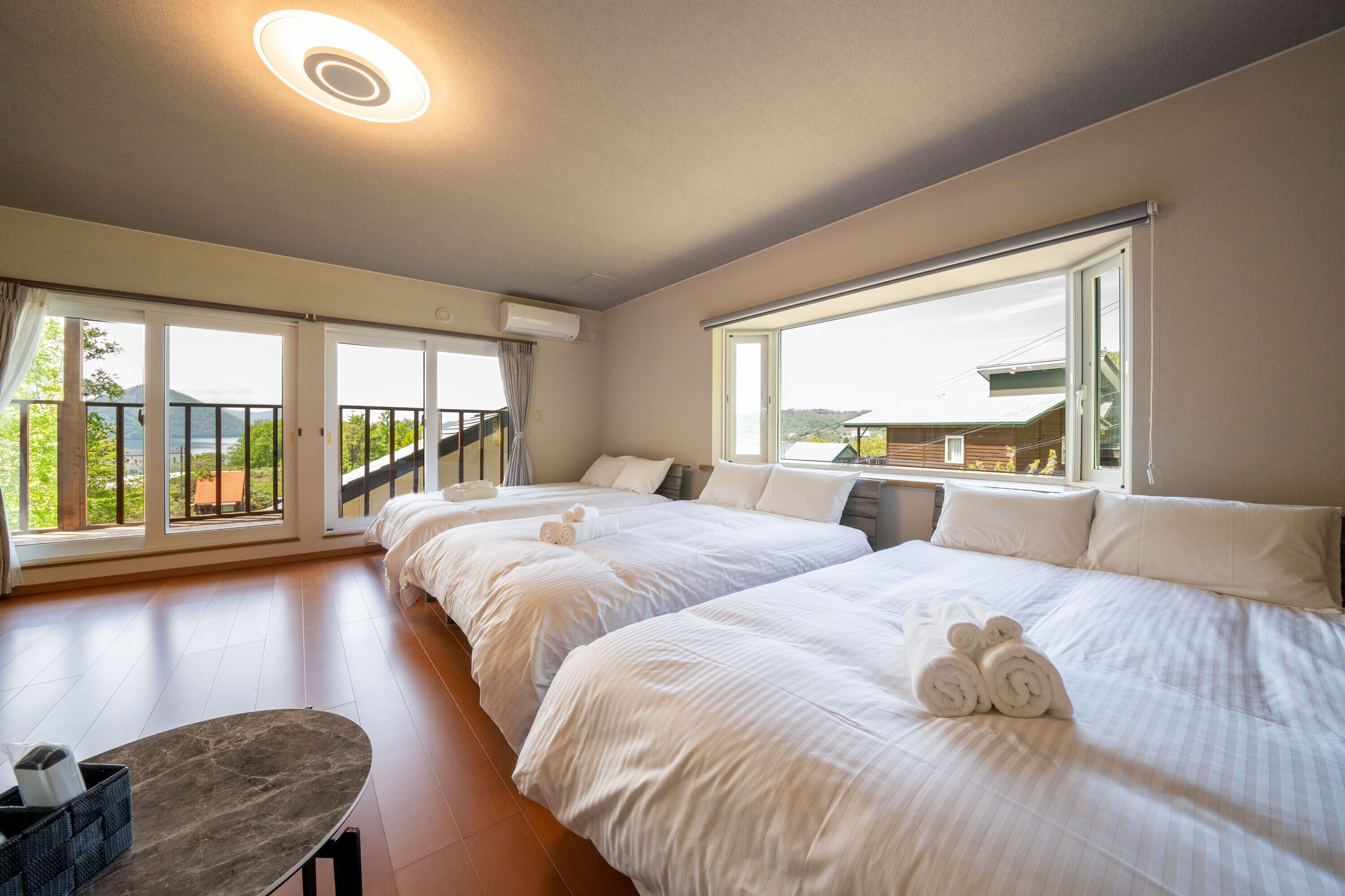 A hotel room with three beds, large windows, a balcony, and a small table.