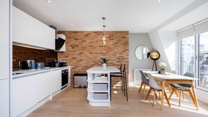 A modern kitchen with white cabinetry, a brick wall, and a dining area with a wooden table and chairs. - Kensington Rooftops, by Veeve (London)