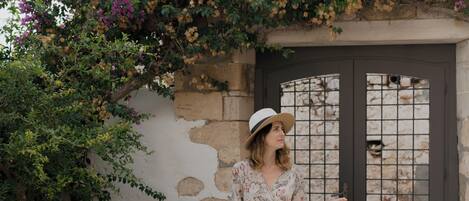 A woman in a floral dress and hat standing in front of a building with a stone wall and a door with a metal grid. There are three large terracotta pots and a smaller pot on the ground.