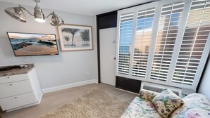 Una sala de estar con alfombra color beige, un sofá estampado con flores, un televisor de pantalla plana, un cuadro enmarcado y una ventana con persianas.