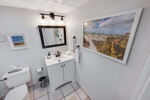 Une salle de bain avec un lavabo blanc, un miroir, un sèche-cheveux et un tableau représentant un paysage, encadré et accroché au mur.