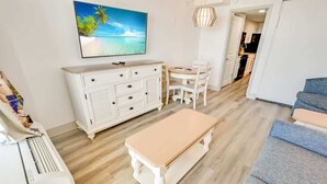 A room with a wooden floor, a white dresser, a TV mounted on the wall, a dining table with chairs, and a sofa.