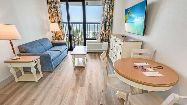 A room with a sofa, a small table, a dining table, chairs, a TV, and a balcony with a view.