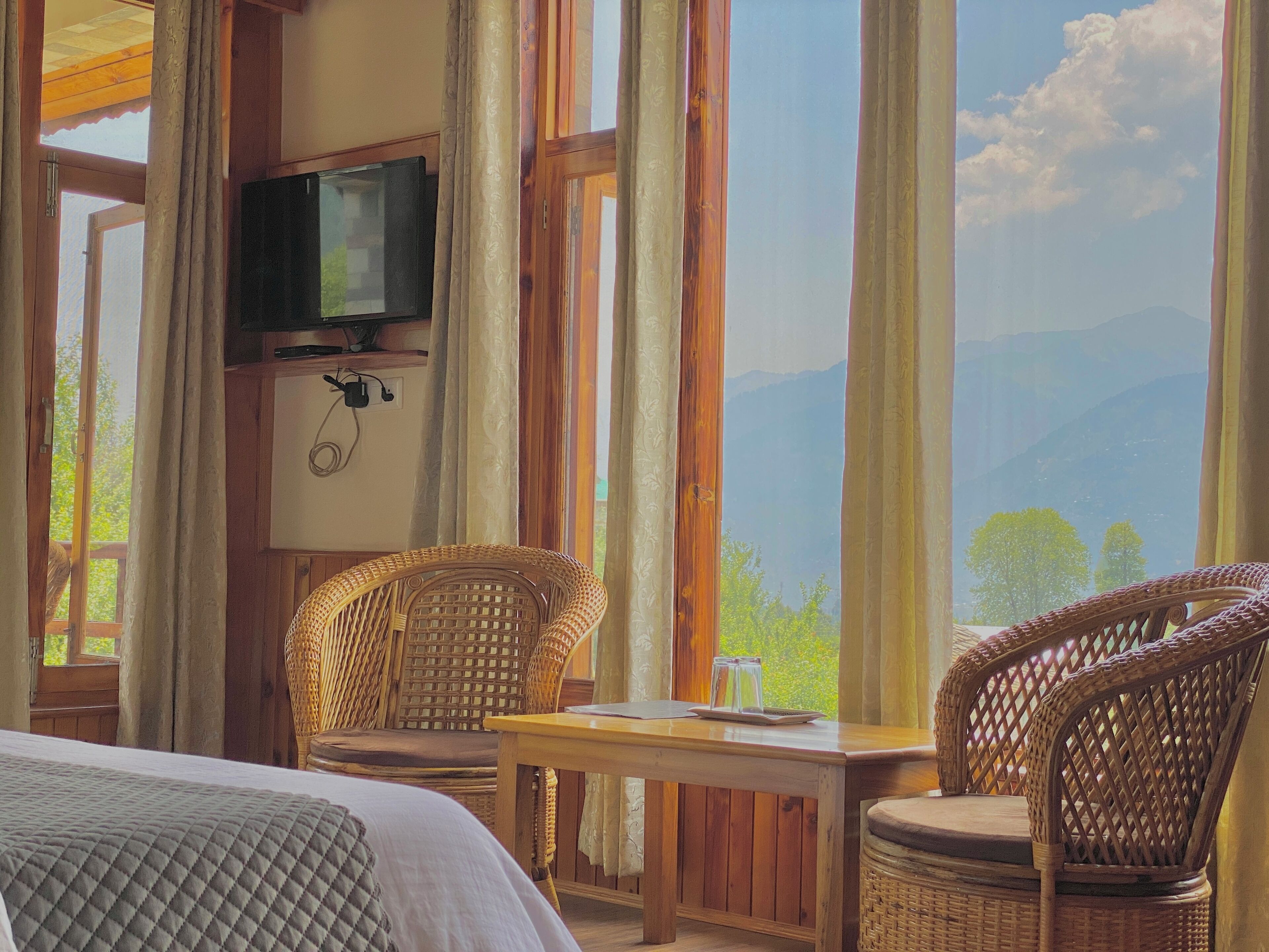 Una habitación con una cama, dos sillones de mimbre, una mesa de madera, un televisor montado en la pared y vistas a la montaña a través de amplios ventanales.
