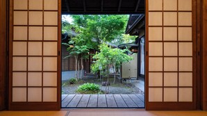 Una habitación japonesa tradicional con suelo de tatami, puertas correderas shoji y vista a un jardín con árboles y una terraza de madera.