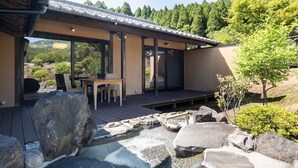 Un edificio de estilo japonés tradicional con un porche de madera, una pequeña fuente termal al aire libre y vistas a los bosques circundantes.
