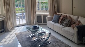Ein Wohnzimmer mit einem Glastisch, einem beigefarbenen Sofa mit gemusterten Kissen und Blick auf Grün hinter einem großen Fenster.