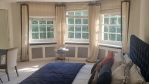 Ein Schlafzimmer mit Bett, Stuhl und einem kleinen Tisch. Es gibt Fenster mit Vorhängen und einen Blick auf Grünflächen.