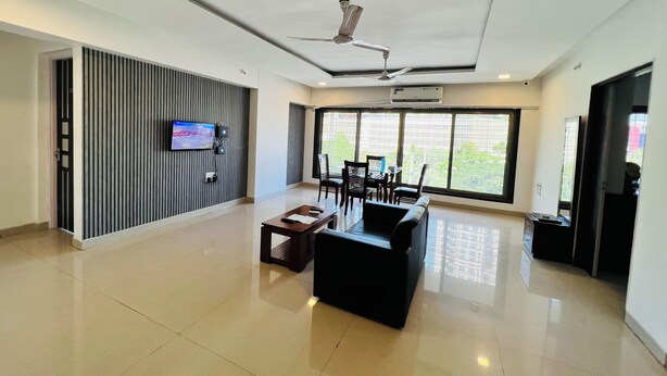 A spacious lobby with a modern design, featuring a flat-screen TV mounted on the wall, a seating area with a sofa and chairs, and a large window offering a view of the outdoors. - Hsquare Service Apartment Malad (Mumbai)