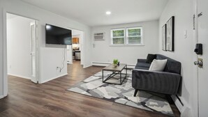 Ein modernes Wohnzimmer mit einem grauen Sofa, einem Couchtisch und einem gemusterten Teppich. Im Hintergrund ist eine Küche mit weißen Schränken und einem an der Wand befestigten, schwarzen Fernseher zu sehen.