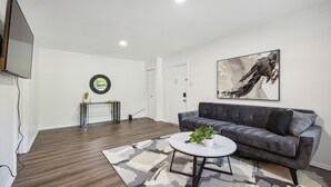Un salon moderne avec un canapé gris, une table basse ronde ornée d’une plante, un grand tableau abstrait et un téléviseur à écran plat fixé au mur.