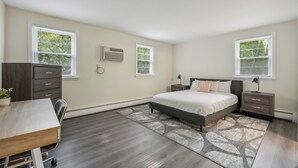 A bedroom with a bed, bedside tables, a desk, and a dresser. There are two windows with blinds, an air conditioning unit, and a phone on the wall. - Pine Brook 1302 I Pool Gym I Free Parking ! (Newark)