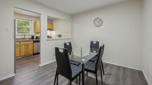Un comedor con una mesa de vidrio y cuatro sillas negras, una cocina con armarios de madera y un reloj de pared.