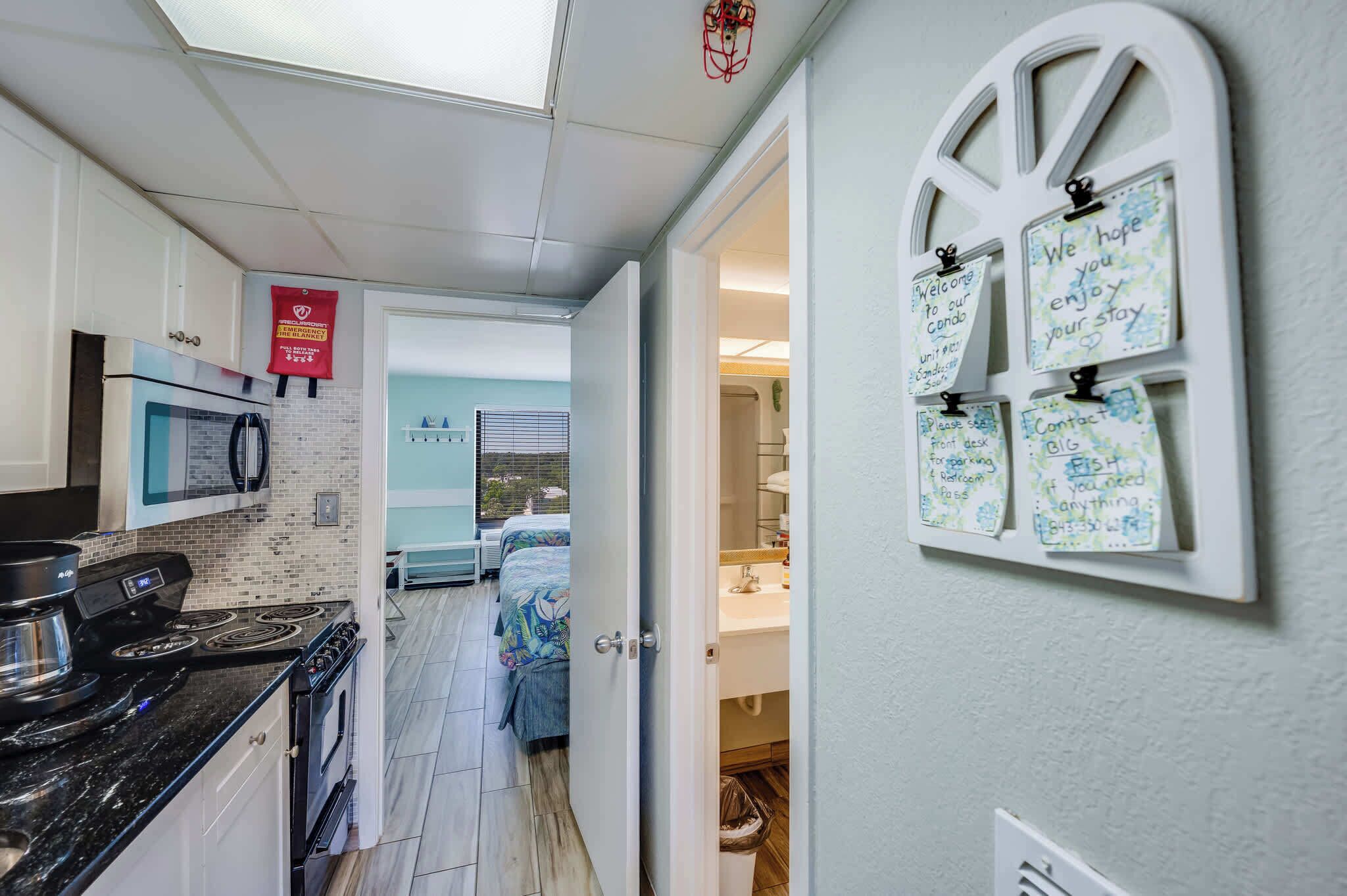 Un couloir étroit, avec une cuisine d’un côté, un lit recouvert d’une courtepointe à motifs floraux, et une salle de bain avec un lavabo et un miroir blancs.