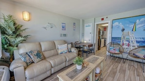 Un salon avec un canapé beige, une table basse, des fauteuils en osier, un tableau représentant une scène de plage et un coin repas avec une table et des chaises.