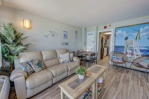 A living room with a beige sofa, a coffee table, wicker chairs, a painting of a beach scene, and a dining area with a table and chairs. - New 1BR Direct Oceanfront, Sandcastle South, Pools (Myrtle Beach)