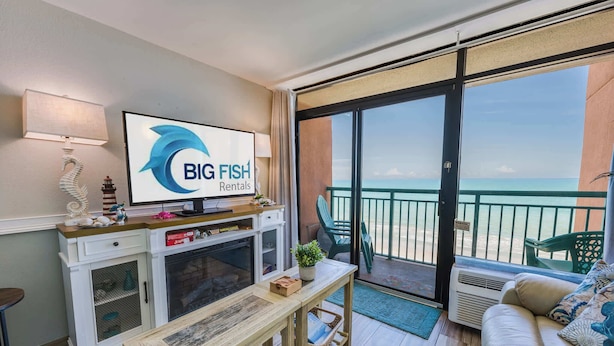 Une chambre dotée d’un téléviseur diffusant des publicités pour « BIG FISH Rentals », d’un balcon offrant une vue sur l’océan, d’un canapé et d’une table à manger.