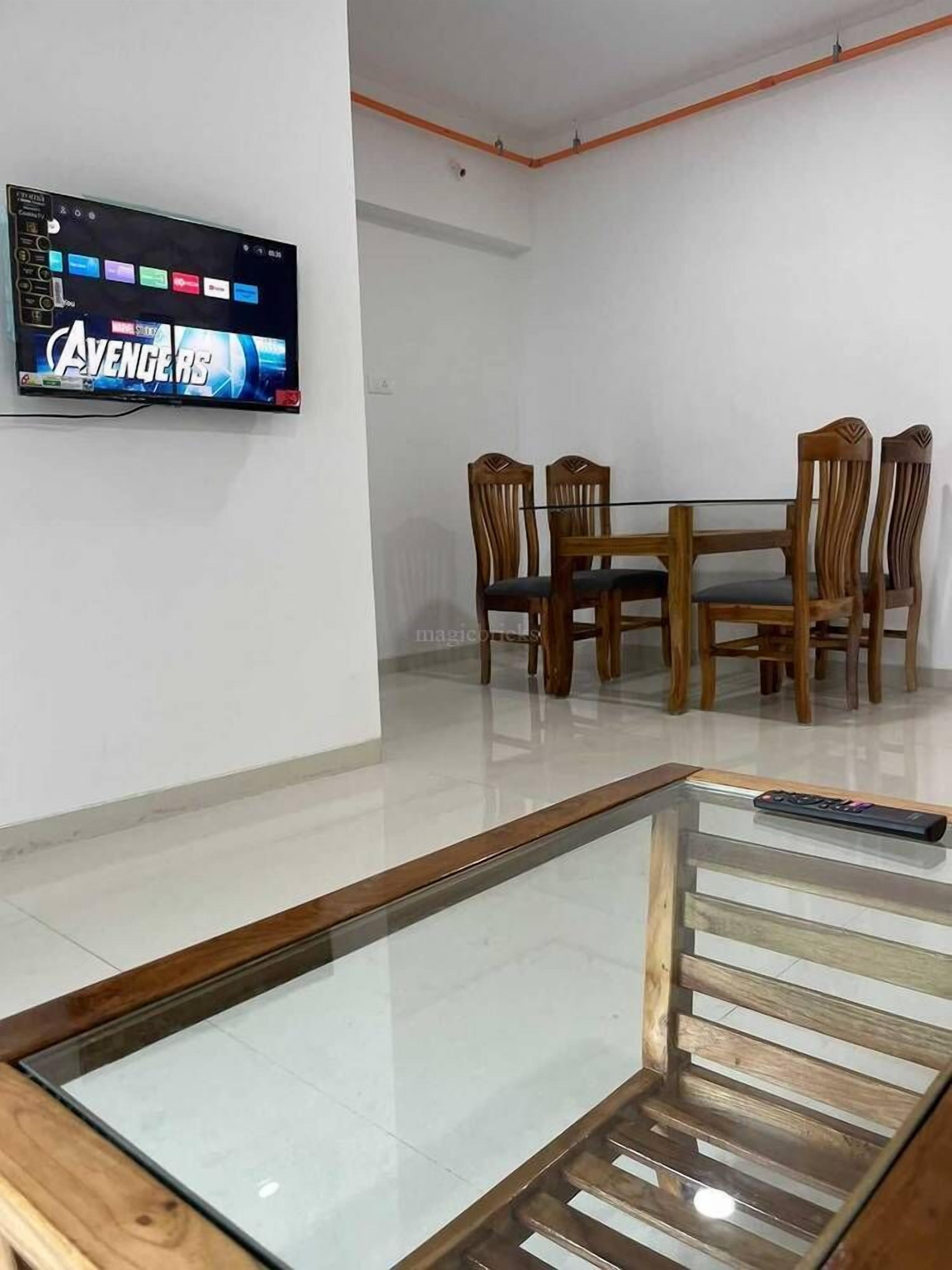 A dining area with a glass-top table and wooden chairs, a mounted flat-screen TV displaying 'AVENGERS 5', and a remote control on the table.