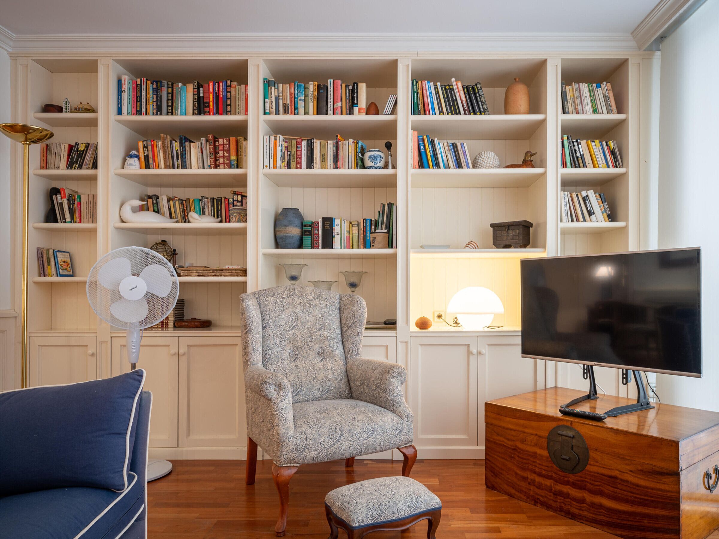 Un salón con un sofá azul, un sillón estampado, una mesa de centro de madera, un televisor y una estantería llena de libros.