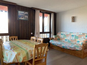 A room with a wooden dining table and chairs, a sofa, a window with curtains, and a framed picture on the wall.