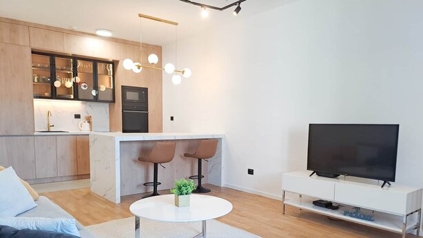A modern living room with a sofa, a coffee table, a TV, and a kitchen area with bar stools. - Lovely Apartment at CITY QUART (Podgorica)
