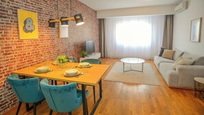 A modern living room with a brick wall, a dining area with a wooden table and blue chairs, and a grey sofa. - Lovely Apartment at CITY QUART (Podgorica)