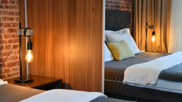 A hotel room with a brick wall, wooden paneling, a bed with white and gray bedding, and two bedside lamps.