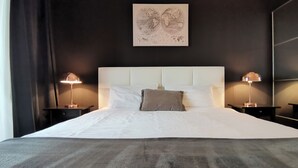 Un lit bien fait, avec du linge de lit blanc et un plaid gris, flanqué de deux tables de chevet surmontées de lampes, dans une pièce aux murs sombres et ornée d’une seule œuvre d’art au-dessus du lit.