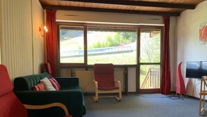 A room with a green sofa, a red armchair, a wooden TV stand, and a large window with a view of greenery and a building. - 3-room Duplex for 6 at slopes, south terrace (Pra-Loup)