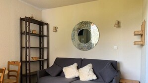 En stue med en grå sofa, to hvide, bløde puder, et træsidebord, en træstol, en træbogreol og et rundt spejl med et geometrisk mønster på væggen.