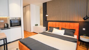 A modern hotel room with a bed, a small kitchenette, a bedside table, and a round table with a chair.