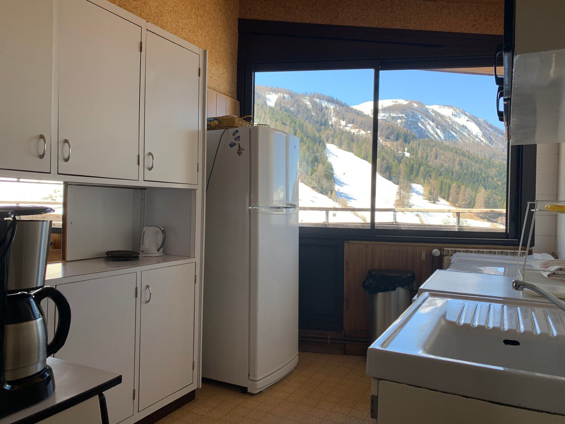 Une cuisine avec des meubles blancs, une bouilloire en acier inoxydable, un réfrigérateur blanc et une vue sur des montagnes enneigées par la fenêtre.