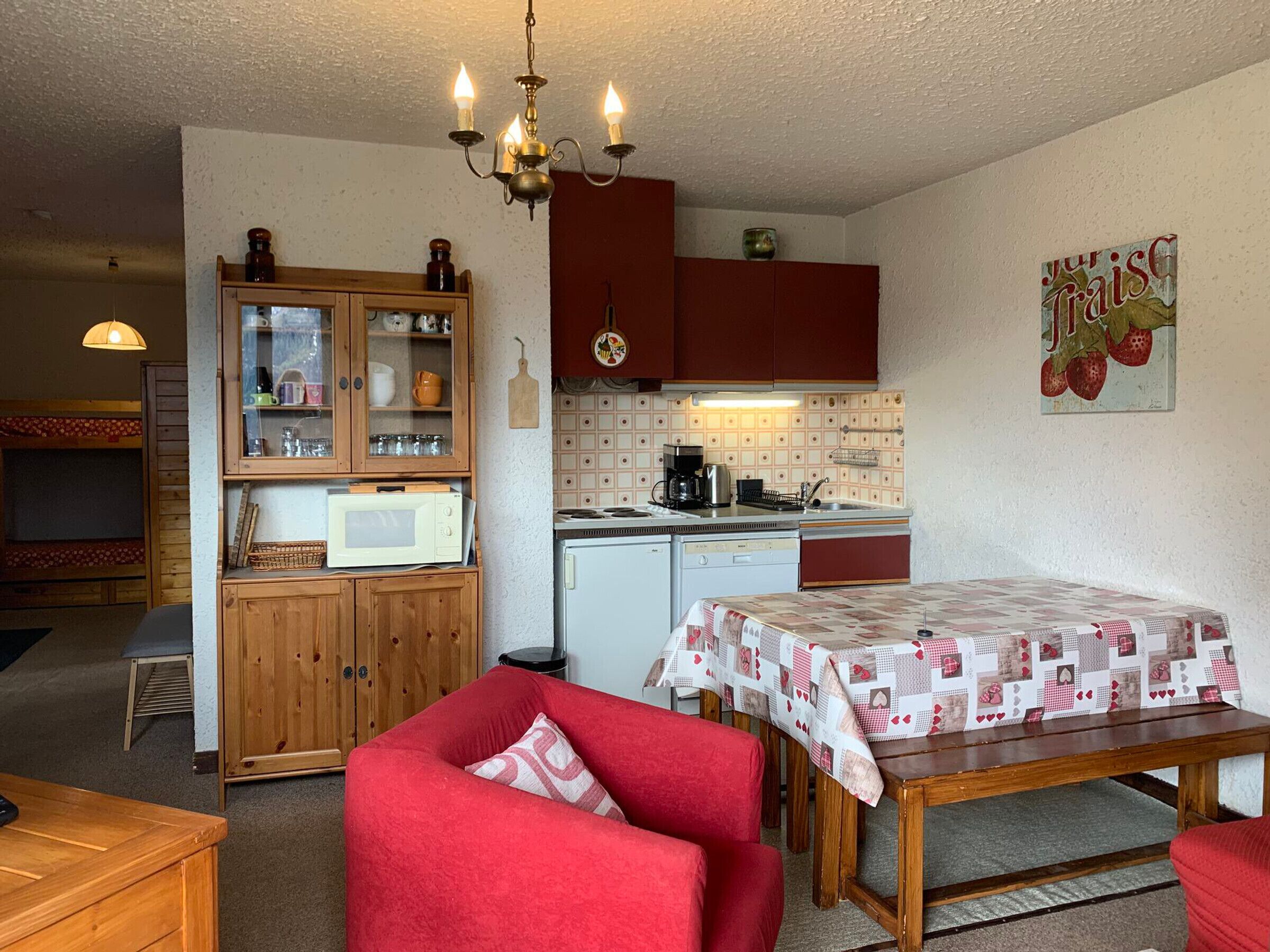 Een kleine keuken met een magnetron, koffiezetapparaat en een spoelbak. Er staat een eettafel met een rode tafelkleed en twee rode stoelen.