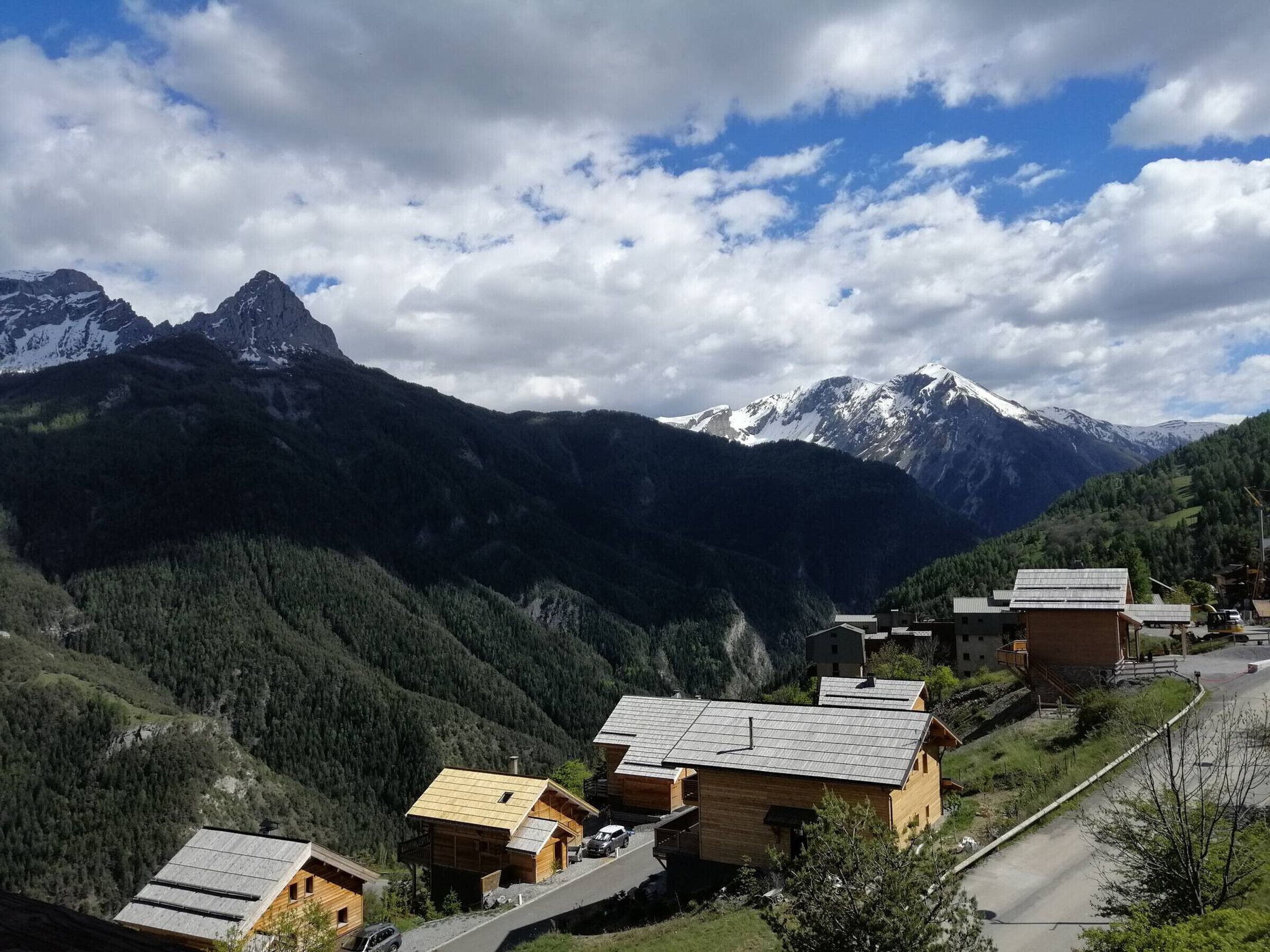 A mountain village with wooden houses, a road, and snow-capped peaks in the background.