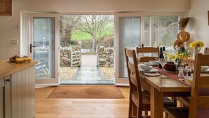 A dining area with a wooden table set for four, a kitchen area with a breakfast setup, and a view of a stone wall and tree through glass doors.