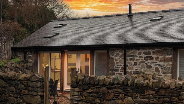 A stone house with a sloped roof, large glass doors, and a stone wall surrounding the property. - Gorse Barn Summary:<br>E (Conwy Principal Area)