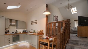 A modern kitchen with a dining area, wooden staircase, and a TV in the living room. - Gorse Barn Summary:<br>E (Conwy Principal Area)