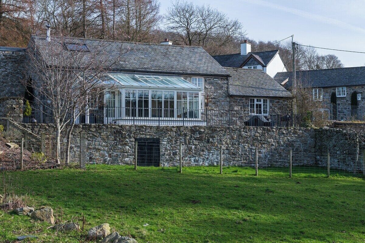A stone house with a large glass conservatory, surrounded by a stone wall and a grassy yard.