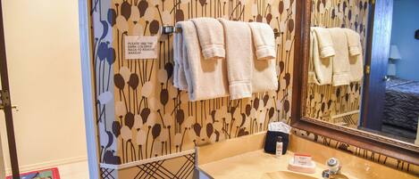 Un baño con un lavabo color beige, un espejo y toallas colgadas en un perchero. Hay un cartel en la pared y un papel pintado con motivos.